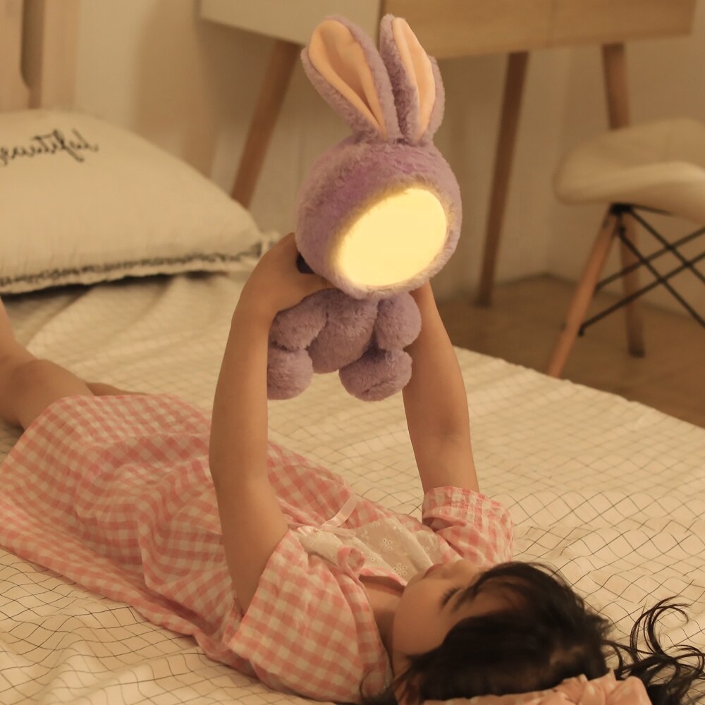 Petite fille qui joue dans son lit avec sa peluche veilleuse en forme de lapin violet 